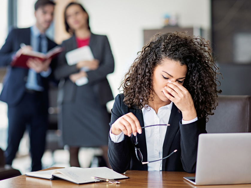 woman in office with head in hands while coworkers gossip behind her