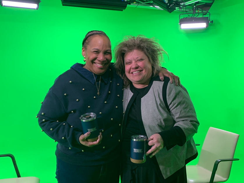 two women standing side by side holding TESU mugs and smiling in front of green screen studio